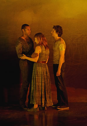 The black man in tight multi-patterend t-shert and jeans is facing the white woman in a long, checkered dress, her sleeveless arms embracing his arms, as the white man in pale t-shirt and dark pants looks at the black man
